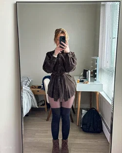 Woman in a brown coat and pink shorts takes a selfie in a bedroom.