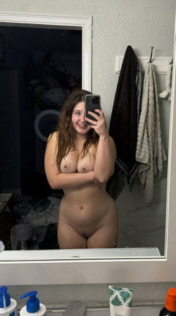 A young woman with long brown hair takes a selfie in a bathroom, smiling and crossing her arms over her chest.