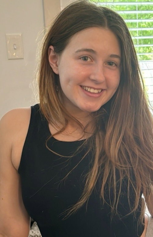 A smiling woman with long brown hair wearing a black tank top indoors.