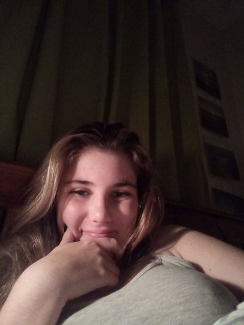 A woman with long blonde hair smiles softly while resting her chin on her hand, wearing a gray tank top in a dimly lit