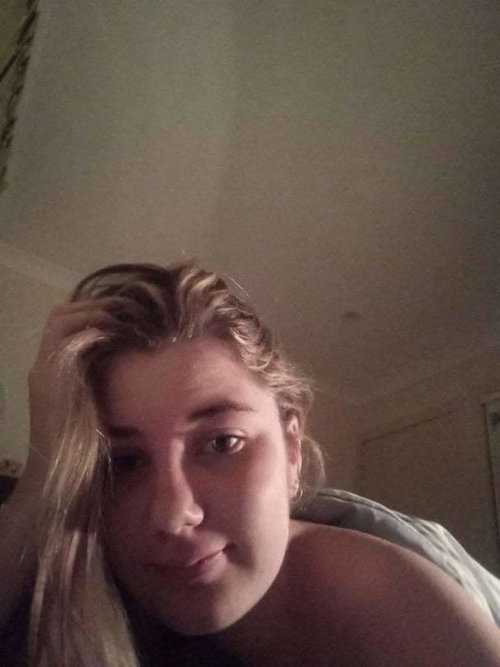 A blonde woman with a curvy build takes a close-up selfie indoors, touching her hair and looking at the camera.