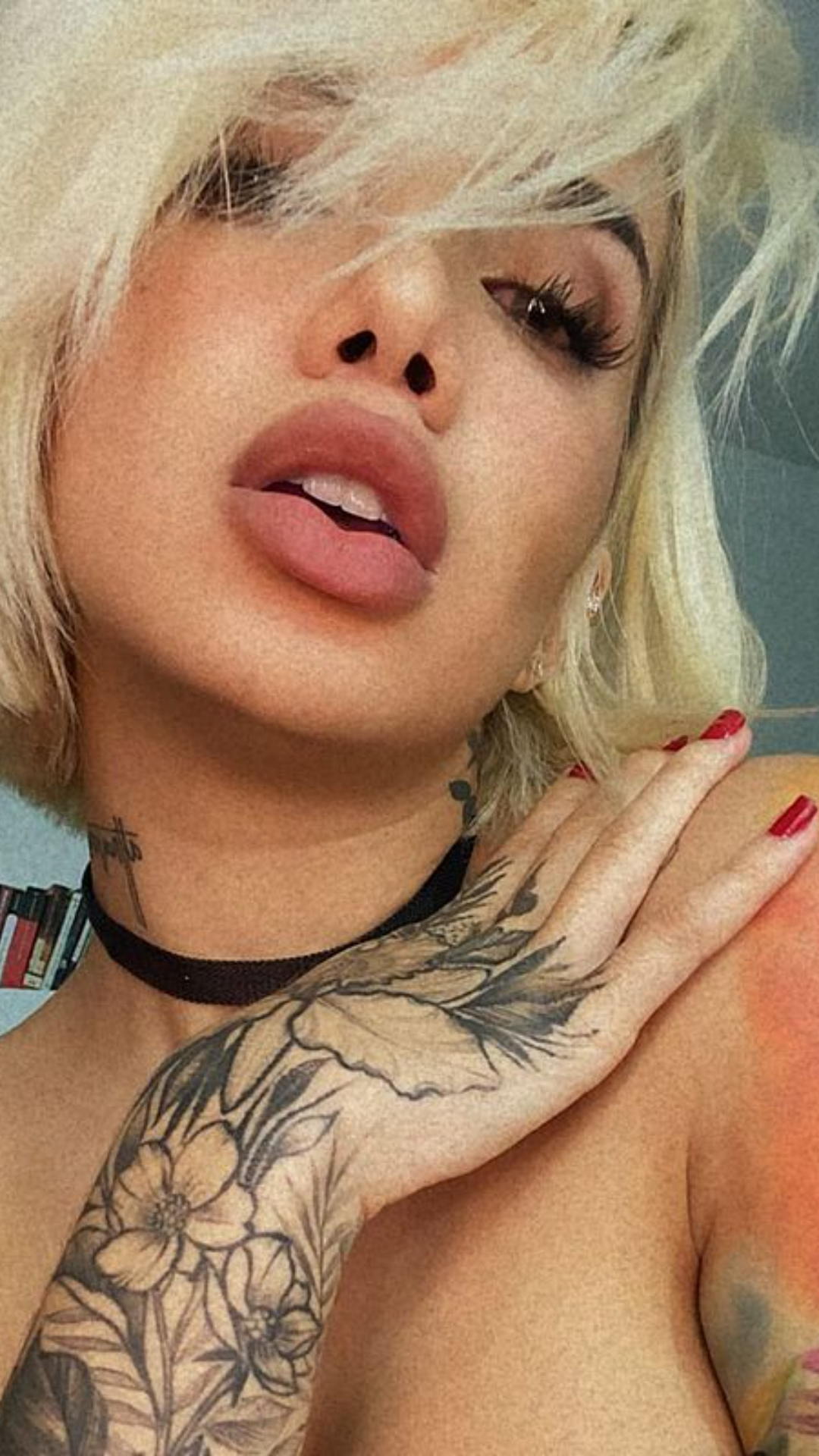 Blonde woman with short hair and floral tattoos on arm and neck, wearing choker, posing topless indoors.