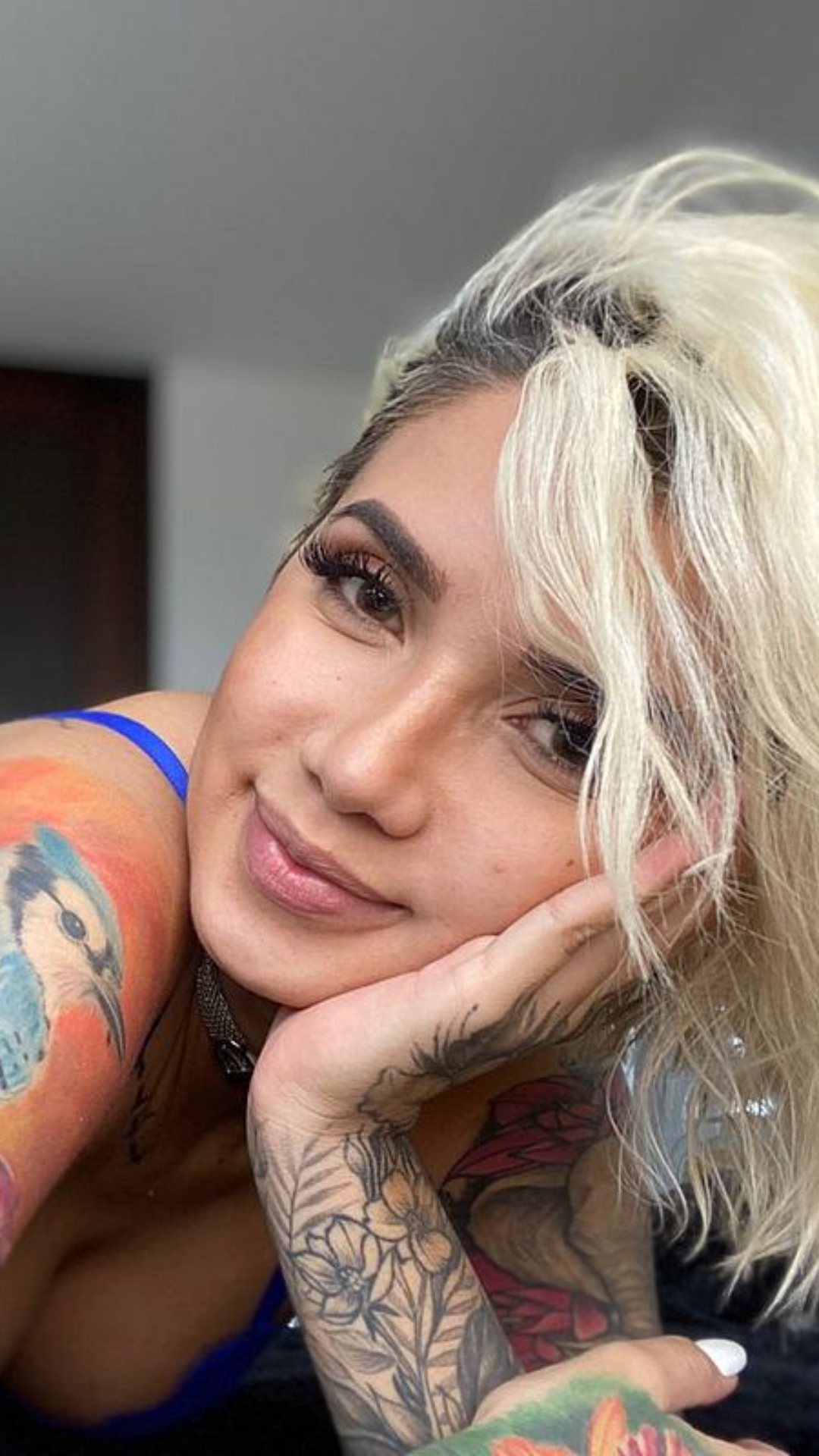 A woman with platinum blonde hair and colorful tattoos on her arms rests her chin on her hand, wearing a blue top