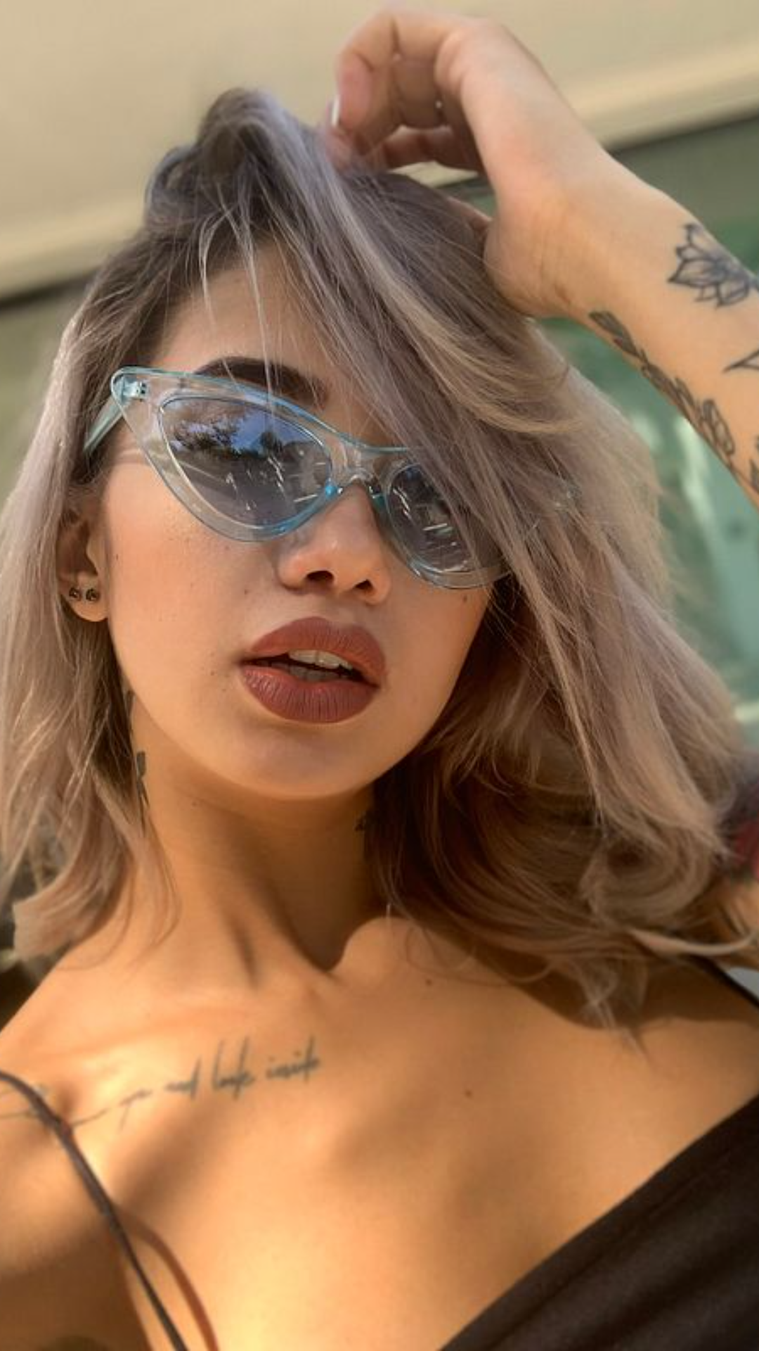 A woman with pastel hair and sunglasses poses outdoors, wearing a black top with visible chest and arm tattoos.
