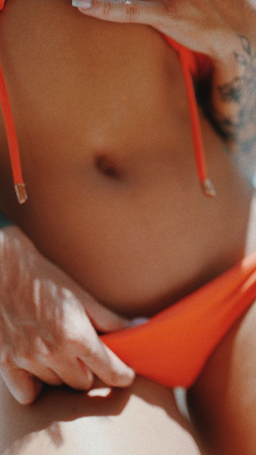 Curvy woman in orange bikini adjusting top, tattoo visible on arm, close-up shot.
