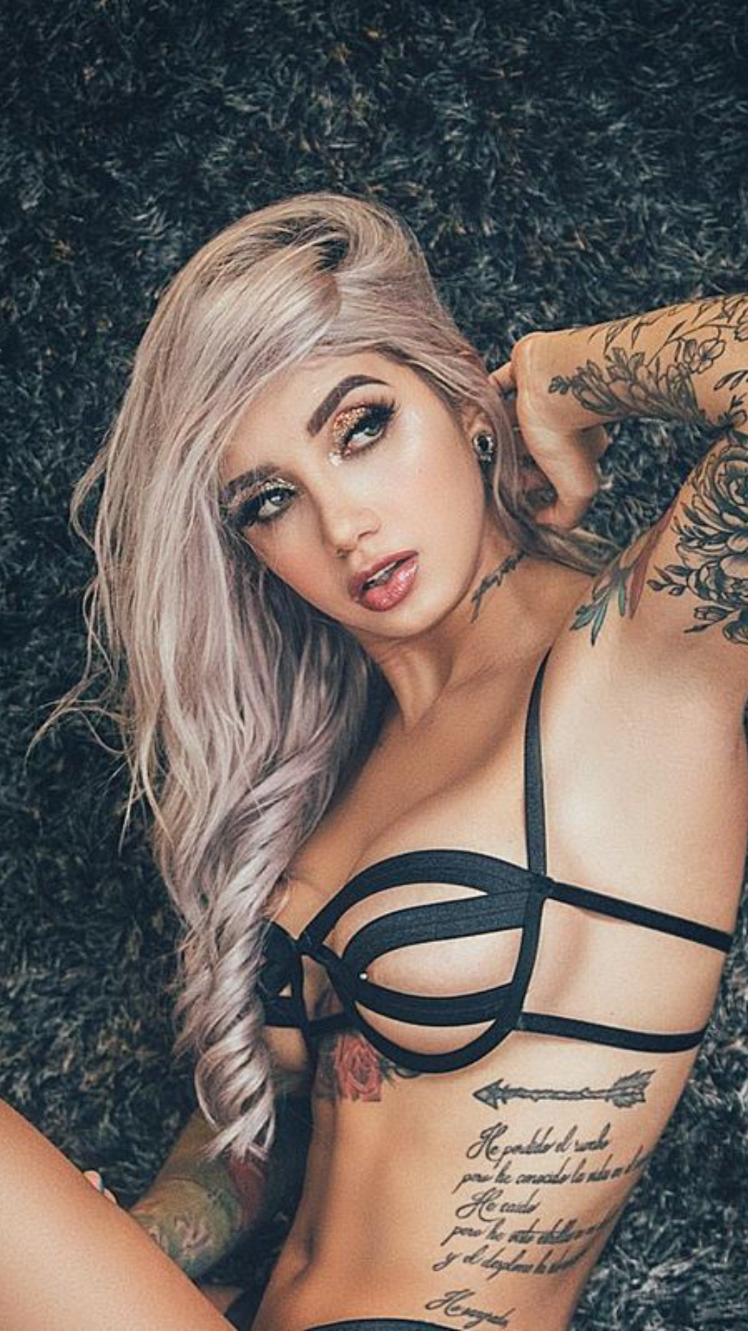 A blonde woman with long wavy hair lies on a dark rug, wearing a black strappy bra, with visible tattoos on her arms