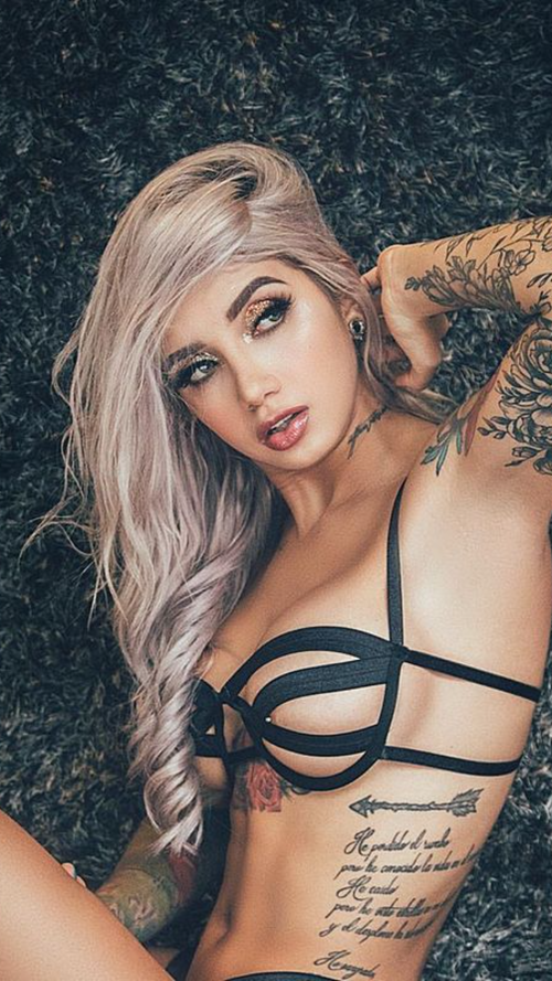 A blonde woman with long wavy hair lies on a dark rug, wearing a black strappy bra, with visible tattoos on her arms