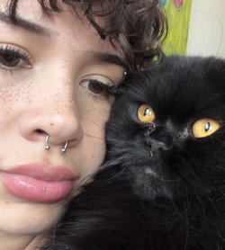 Person with dark curly hair and a nose ring cuddles a black cat with yellow eyes.
