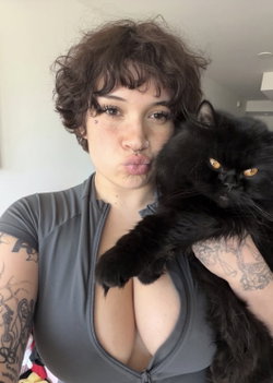 Person with curly hair and tattoos holding a black cat, wearing a gray top.