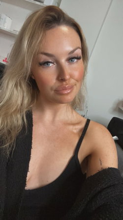 Blonde woman with blue eyes and freckles wearing a black tank top and cardigan, taking a selfie indoors.