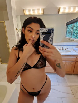 Brunette woman in black mesh lingerie takes a mirror selfie in a bright bathroom, touching her nose with one hand.