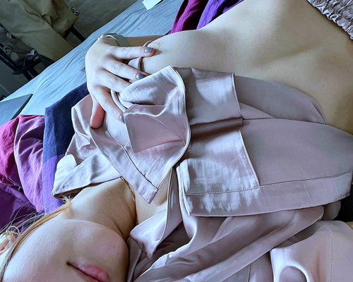 A blonde woman in an unbuttoned pink satin top lies on purple bed sheets.