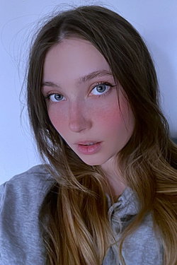 A young woman with long brown hair and freckles poses in a grey hoodie for a selfie.