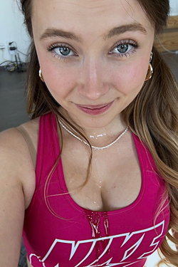 Close-up of a woman with brown hair and blue eyes, wearing a pink Nike tank top, smiling at the camera in a casual