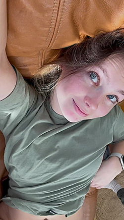 A person with light skin and blue eyes lies on a brown leather couch wearing a green shirt.