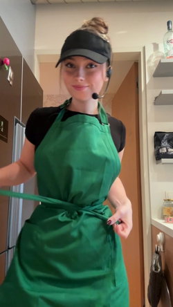 A woman in a green apron and black cap stands in a kitchen.