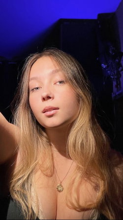 Blonde woman with long hair taking a selfie in dim blue-lit room, wearing lace top and gold pendant necklace.