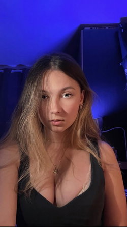 A woman with long brown hair and a black top poses in a dimly lit room with blue ambient lighting.