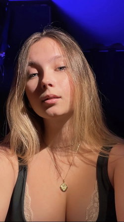 Blonde woman with shoulder-length hair wearing a black lace-trimmed top, posing in dim blue-lit setting.