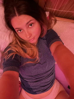 A woman with dark hair and highlights lies on a bed, wearing a gray crop top and pink bottoms, taking a selfie in warm