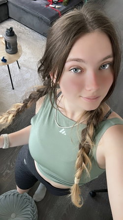A young woman with braided hair and blue eyes wears a green crop top and black leggings, taking a selfie in a modern