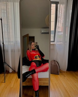 Blonde woman in sheer red Elastigirl cosplay bodysuit, black gloves and boots, mirror selfie on bed.
