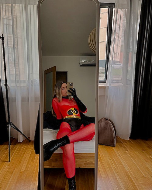 Blonde woman in sheer red Elastigirl cosplay bodysuit, black gloves and boots, mirror selfie on bed.