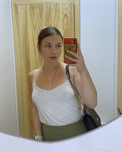 A woman with dark hair takes a mirror selfie in a polka-dot tank top and olive green skirt, holding a red phone with a