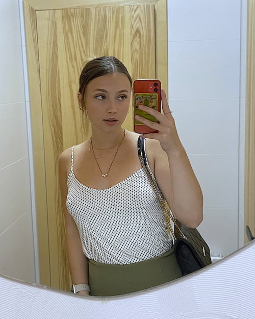 A woman with dark hair takes a mirror selfie in a polka-dot tank top and olive green skirt, holding a red phone with a