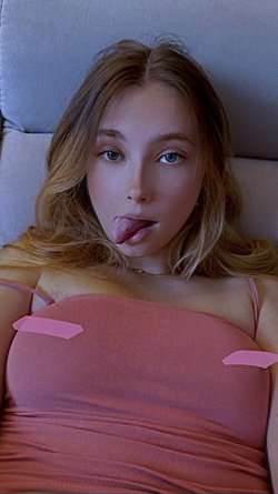 Blonde woman with wavy hair lies on a couch, wearing a pink top, sticking her tongue out playfully at the camera.
