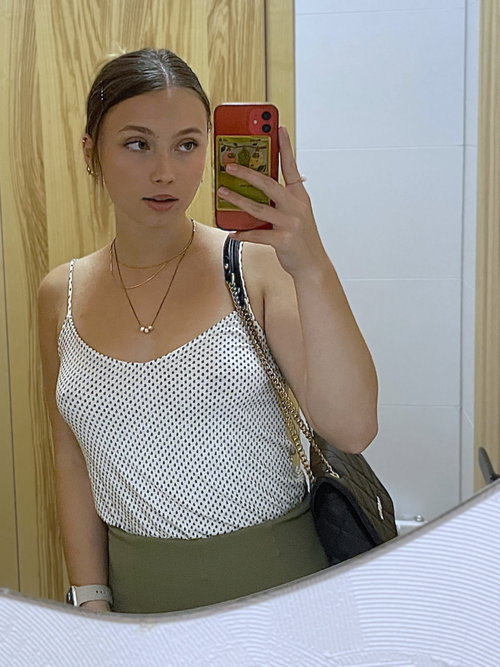 A young woman with dark hair takes a mirror selfie in a polka-dot tank top and olive green skirt, holding a red phone