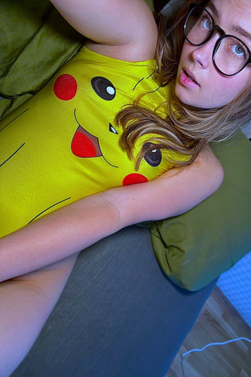 A woman with glasses and light brown hair lies on a green cushion wearing a yellow Pikachu tank top, looking up at the