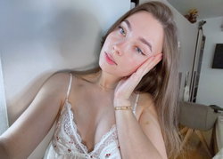 A woman with long brown hair and blue eyes wears a white lace camisole, leaning against a wall with one hand on her