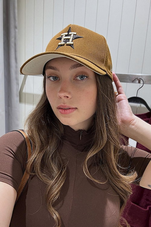 A young woman with long wavy brown hair and blue eyes wears a brown baseball cap and a brown zip-up top, posing indoors