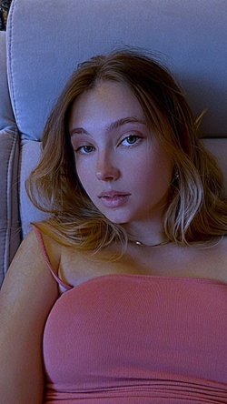 A young woman with long brown hair lies on a couch wearing a pink top.