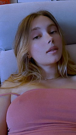 Blonde woman with nose piercings lies on a bed, wearing a pink strapless top, looking directly at the camera.