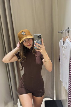Brunette woman in a brown zip-up bodysuit and gold cap takes a mirror selfie in a room with clothes hanging nearby.