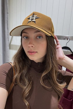 A young woman with long wavy brown hair and blue eyes wears a brown baseball cap and a brown zip-up top, posing indoors