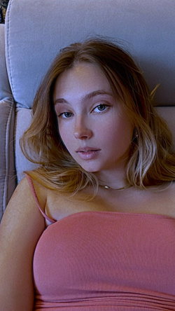 A woman with wavy light brown hair reclines on a couch wearing a pink top.