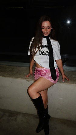 Brunette woman in a white crop tee, pink sequined mini skirt, and black thigh-high boots, posing on a concrete ledge at