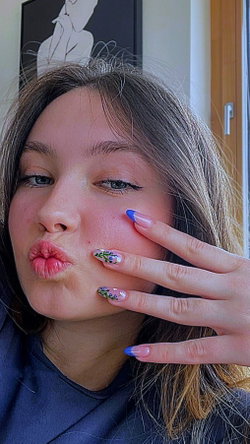 A woman with brown hair and floral nail art puckers her lips while holding her hand near her face, wearing a dark top