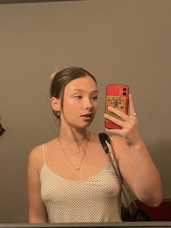 A young woman with dark hair in a high ponytail takes a mirror selfie, wearing a white polka-dot tank top and layered