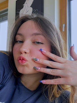A young woman with light brown hair and floral nail art makes a playful kiss face in a selfie, wearing a dark blue top