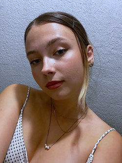 A young woman with brown hair and subtle makeup wears a patterned spaghetti strap top, posing against a textured wall