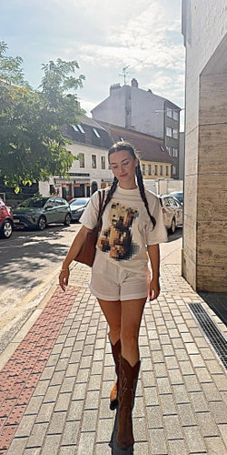 A woman with braided hair walks down a cobblestone street wearing a white t-shirt, shorts, and tall boots.