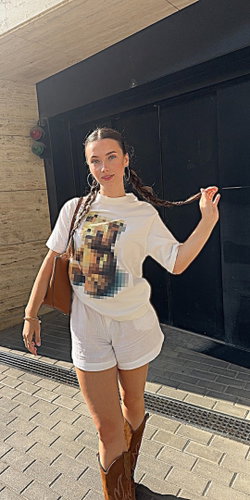 A woman with braided hair wearing a white t-shirt and shorts stands on a sidewalk.