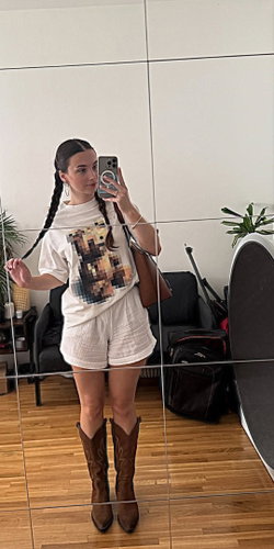Woman with long braided hair in white outfit with brown boots taking a selfie in a mirror.