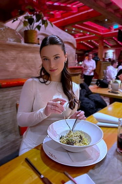 A brunette woman in a white dress sits at a restaurant table eating pasta.