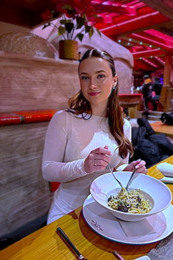 A woman with long brown hair and a white top eats pasta in a cozy restaurant.