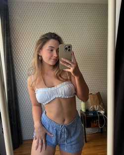 A woman with long blonde hair in a white crop top and blue shorts takes a selfie in a bathroom.